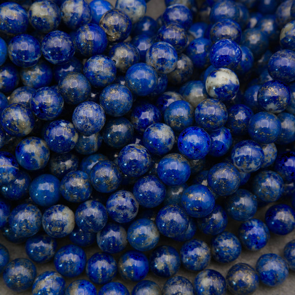 A close-up pile of polished round beads made of lapis lazuli, showing deep blue and white marbled patterns with tiny golden flecks.