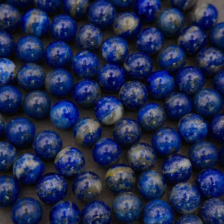 A close-up pile of polished spherical beads in lapis lazuli, showing deep blue with white marbling and subtle golden flecks, resting on a dark surface.