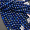A close-up pile of round blue gemstone beads with white cloudy inclusions, photographed on a gray surface with product tags visible.