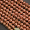 Close-up of copper-colored metallic round beads strung on a cord, showing a hammered texture and warm rose-gold/copper tone against a dark background.