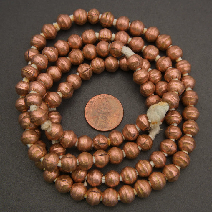 A coiled strand of metallic copper-colored beads with a small copper penny in the center for scale, photographed on a dark gray background.