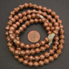 A coiled strand of metallic copper-colored beads with a small copper penny in the center for scale, photographed on a dark gray background.