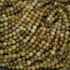 A close-up pile of small faceted round beads in earthy olive green, tan, brown, and cream tones on a dark gray background.