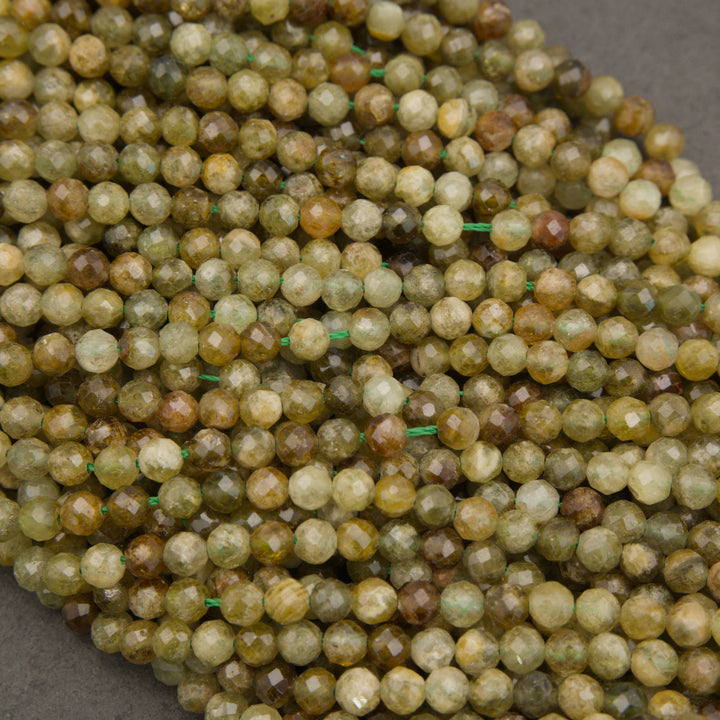 A close-up pile of faceted round beads in earthy olive green, tan, brown, and cream tones, strung on thin green cords against a dark gray background.