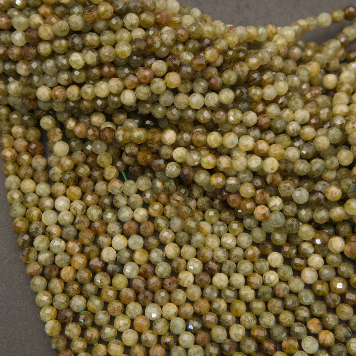 A close-up pile of small faceted round beads in earthy olive green, tan, brown, and cream tones on a dark gray background.
