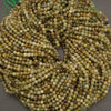 A close-up pile of small faceted round beads in earthy olive green, tan, brown, and cream tones on a dark gray background.