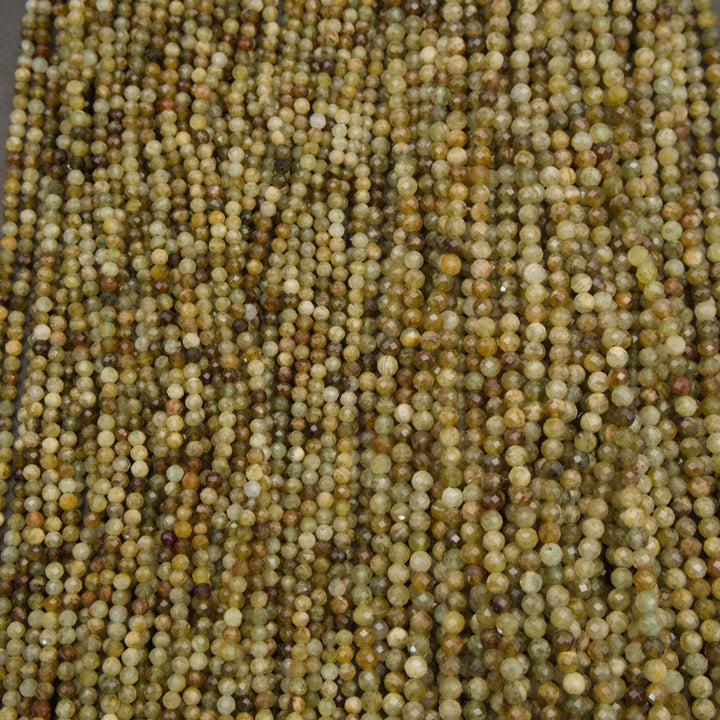 A close-up pile of small faceted round beads in earthy olive green, tan, brown, and cream tones on a dark gray background.