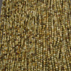 A close-up pile of small faceted round beads in earthy olive green, tan, brown, and cream tones on a dark gray background.