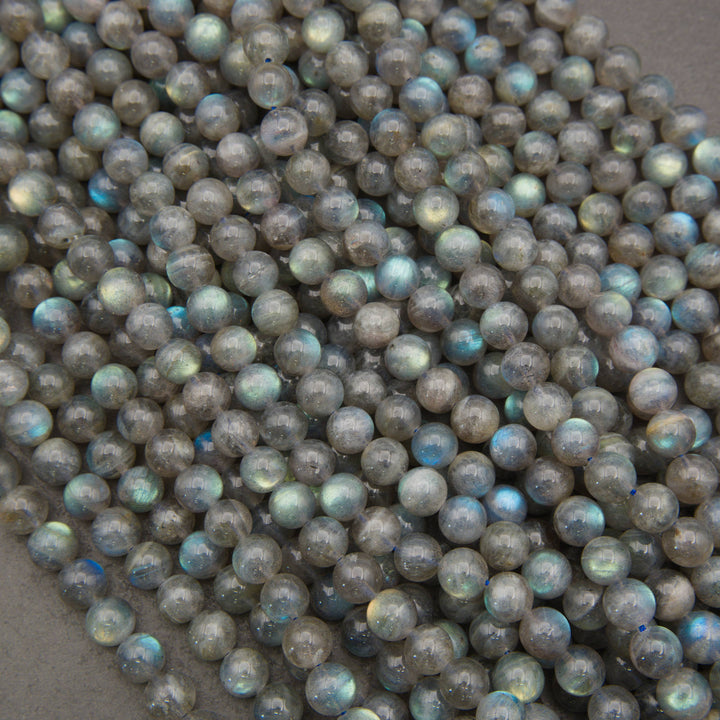A pile of small, round, polished labradorite beads with a gray base and a shimmering, iridescent blue-green finish, resting on a neutral gray surface.