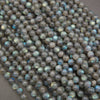 A pile of small, round, polished labradorite beads with a gray base and a shimmering, iridescent blue-green finish, resting on a neutral gray surface.