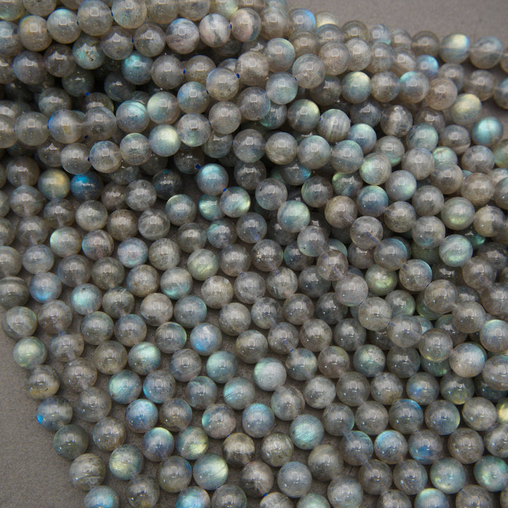 A pile of small, round, faceted beads made from labradorite stone, showing a characteristic gray base color with iridescent flashes of blue, green, and gold.