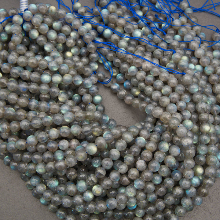 Multiple strands of round labradorite beads with a gray base and iridescent blue-green flashes, strung on blue cords and piled on a neutral surface.
