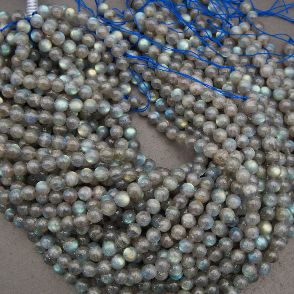 Multiple strands of round labradorite beads with a gray base and iridescent blue-green flashes, strung on blue cords and piled on a neutral surface.