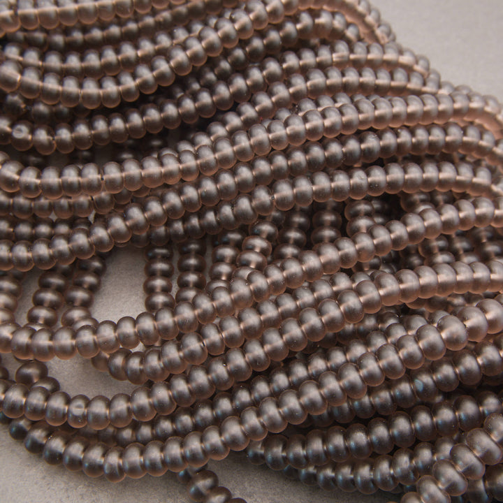 Close-up of multiple strands of small, round, metallic bronze beads arranged diagonally on a light neutral background.