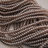 Close-up of multiple strands of small, round, metallic bronze beads arranged diagonally on a light neutral background.