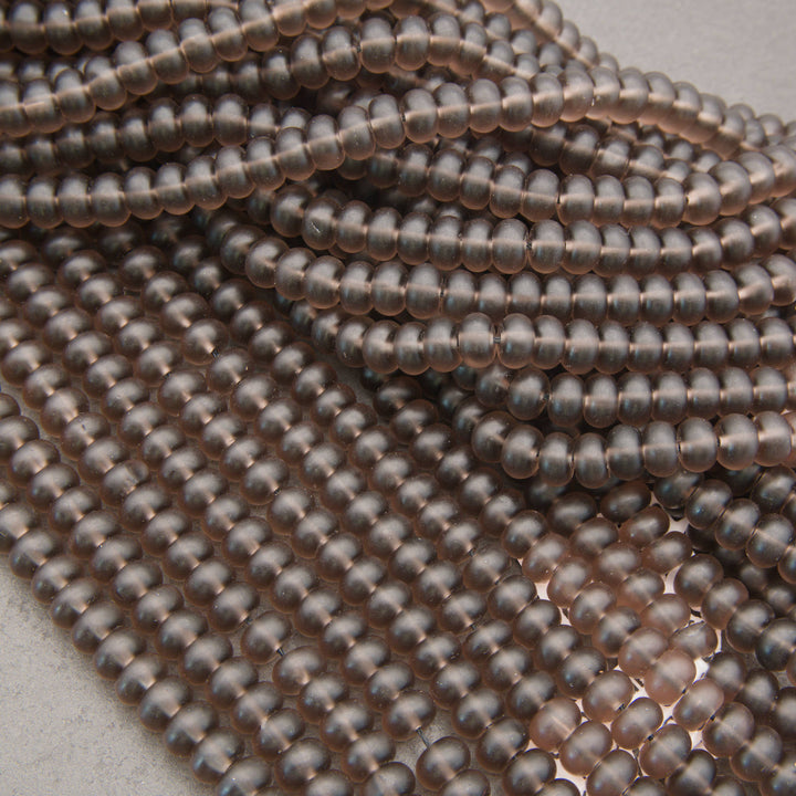 Close-up of multiple strands of small, smooth, metallic oval beads in a warm coppery rose-gold tone, arranged on a light neutral surface.