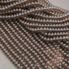 Close-up of multiple strands of small, smooth, metallic oval beads in a warm coppery rose-gold tone, arranged on a light neutral surface.
