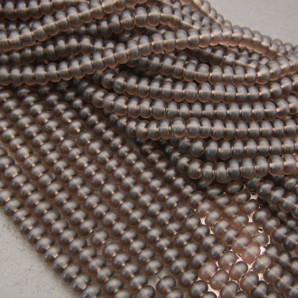 Close-up of multiple strands of small, smooth, metallic oval beads in a warm coppery rose-gold tone, arranged on a light neutral surface.