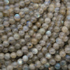 A close-up pile of round labradorite gemstone beads showing a cloudy, semi-translucent gray and beige appearance with subtle blue iridescent flashes.