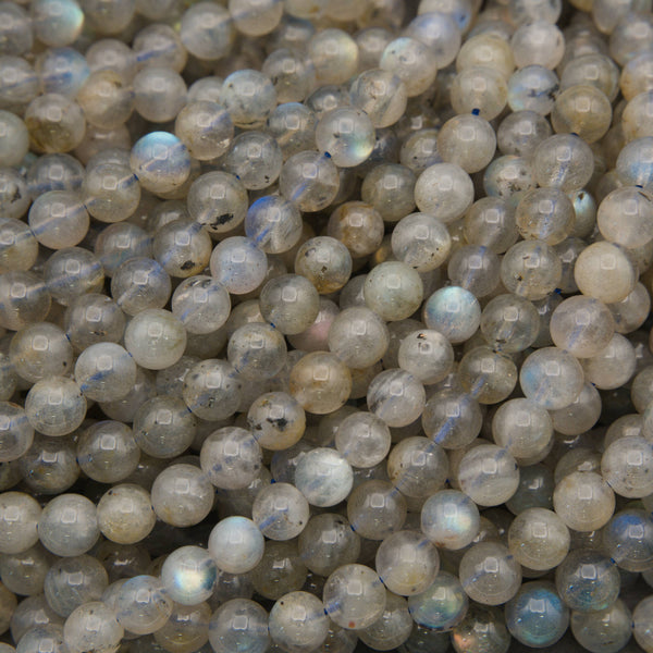 A close-up pile of round labradorite gemstone beads showing a cloudy, semi-translucent gray and beige appearance with subtle blue iridescent flashes.