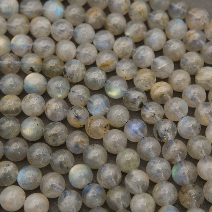 A close-up pile of round, polished labradorite beads showing a milky white to light gray base with blue and green iridescent flashes.
