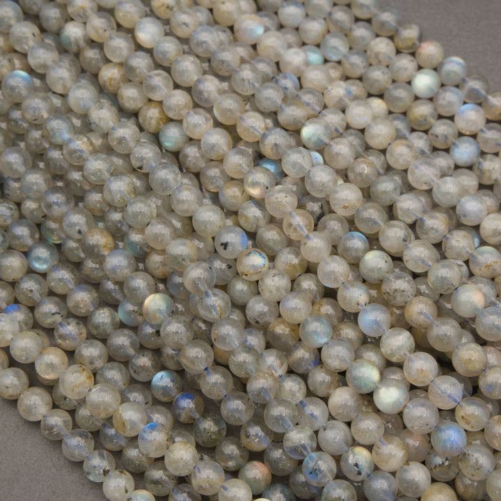 A pile of small, round, polished labradorite gemstone beads with a natural gray and beige base and a subtle iridescent blue flash.