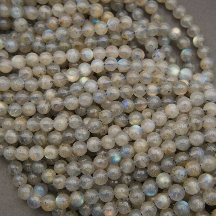 A pile of round labradorite beads with a milky white to light gray base and a subtle iridescent blue-green flash, strung together on thin cords.