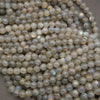 A pile of round labradorite beads with a milky white to light gray base and a subtle iridescent blue-green flash, strung together on thin cords.