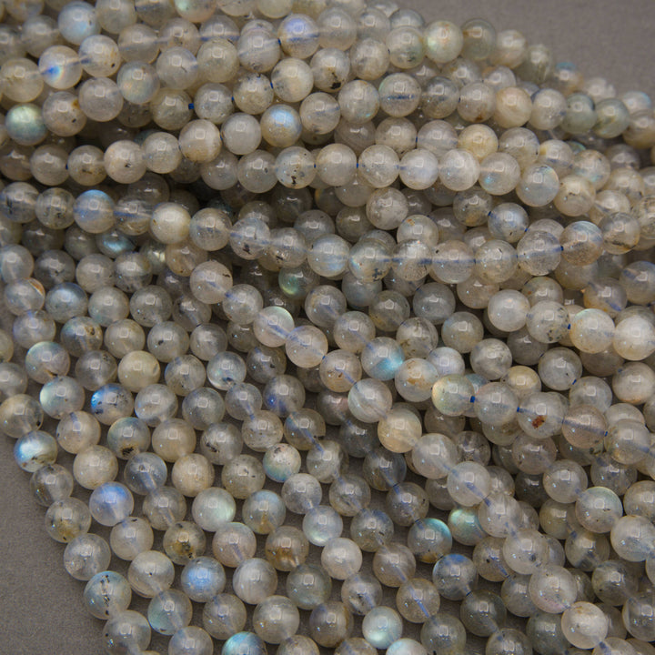 A close-up pile of faceted round beads with a milky white to light gray base and subtle iridescent blue, green, and violet flashes, photographed against a neutral gray background.