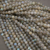 A close-up pile of faceted round beads with a milky white to light gray base and subtle iridescent blue, green, and violet flashes, photographed against a neutral gray background.