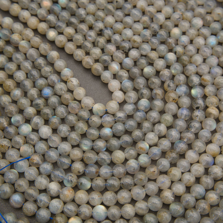 Close-up of many small round labradorite beads in a light gray, smoky translucent stone with subtle iridescent blue flashes, arranged on a neutral surface.