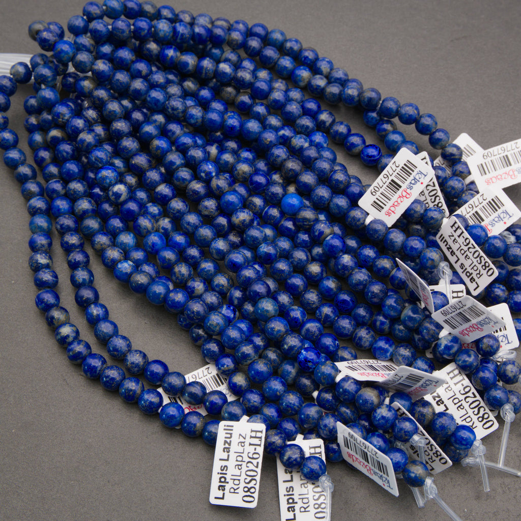 A close-up pile of round, polished lapis lazuli stone beads in deep blue with white cloudy inclusions, resting on a neutral gray surface.