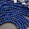 Loose round lapis lazuli stone beads in deep blue with white and gray matrix, arranged in curved strands on a gray surface.