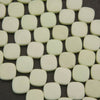 A close-up of many small rounded square tiles in a warm off-white or cream color, arranged on a dark background.