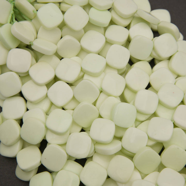 A close-up pile of small, rounded, pale mint-green square-shaped beads or decorative pieces on a dark background.
