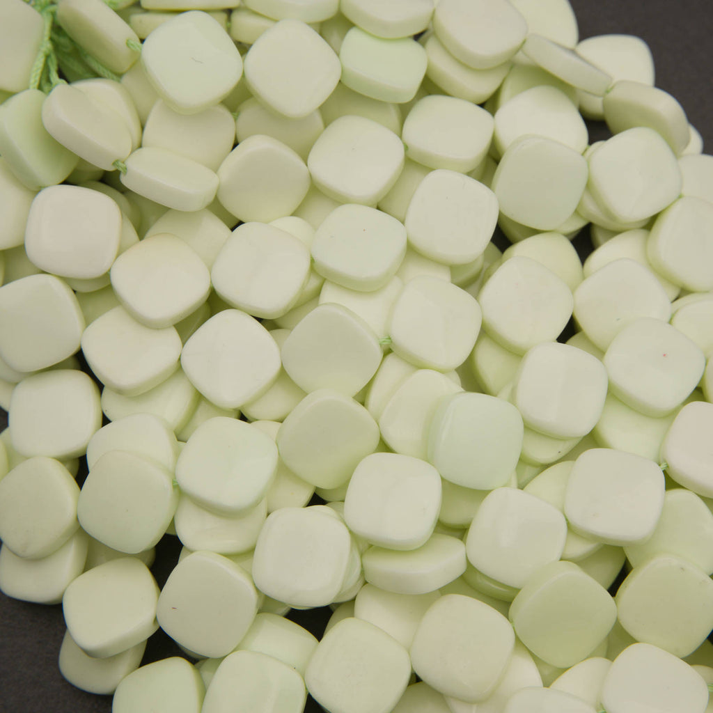 A close-up pile of small, rounded, pale mint-green square-shaped beads or decorative pieces on a dark background.