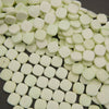 A pile of small, rounded square beads in a pale mint green color, arranged on a dark gray background.