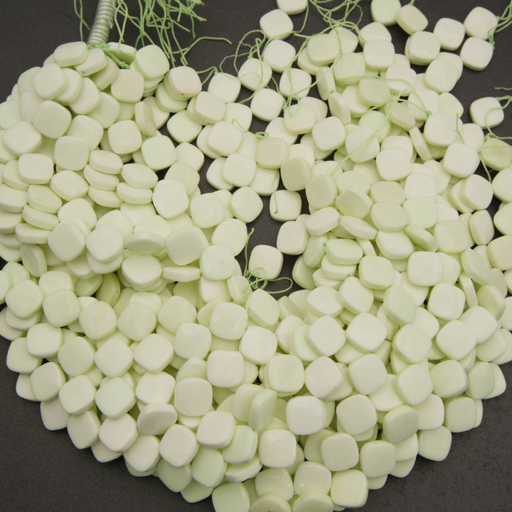 A pile of small, pale greenish-white square beads with rounded edges, strung on thin green cords, displayed on a dark gray background.