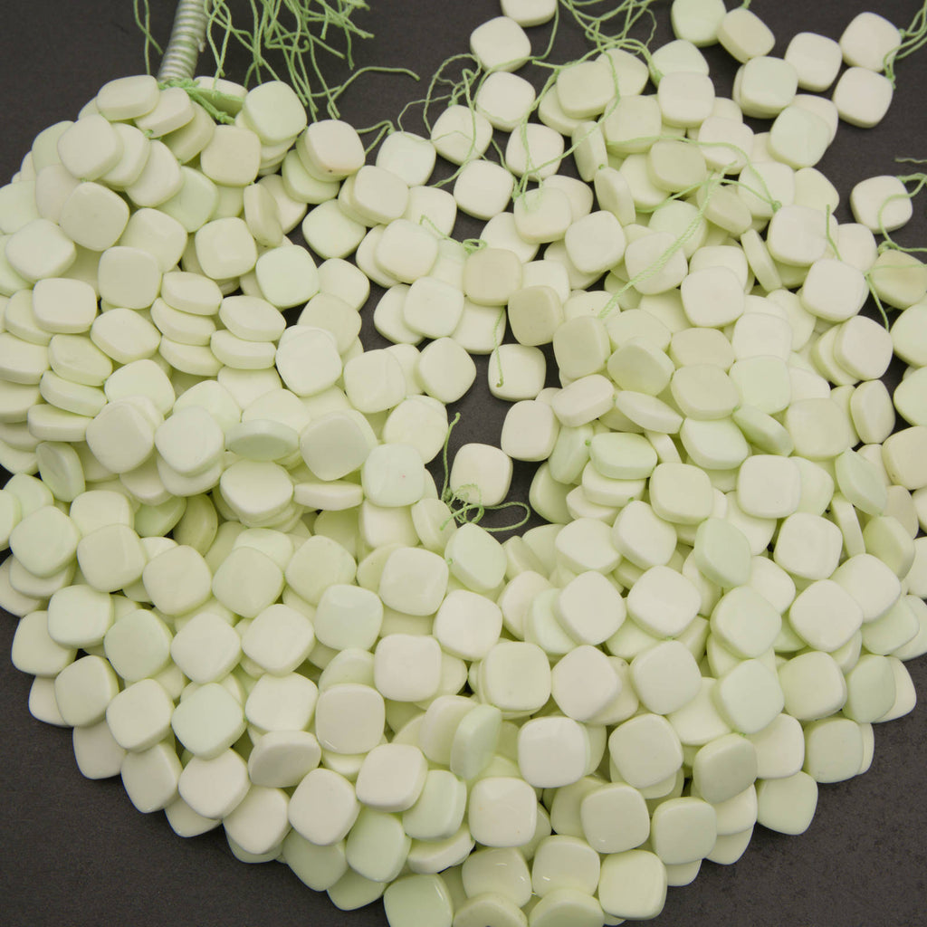 A pile of small, pale greenish-white square beads with rounded edges, strung on thin green cords, displayed on a dark gray background.