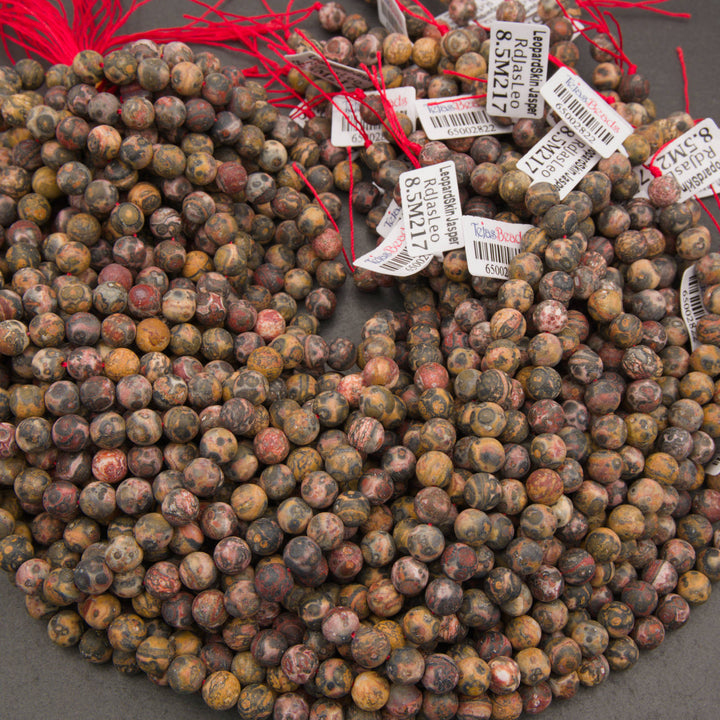 A large pile of round gemstone beads in earthy brown, tan, gray, and muted red tones, each tied with a red string and labeled with white tags.