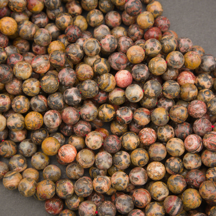 A pile of round gemstone beads in earthy, mottled colors including brown, yellow, red, and gray, arranged on a dark gray surface.