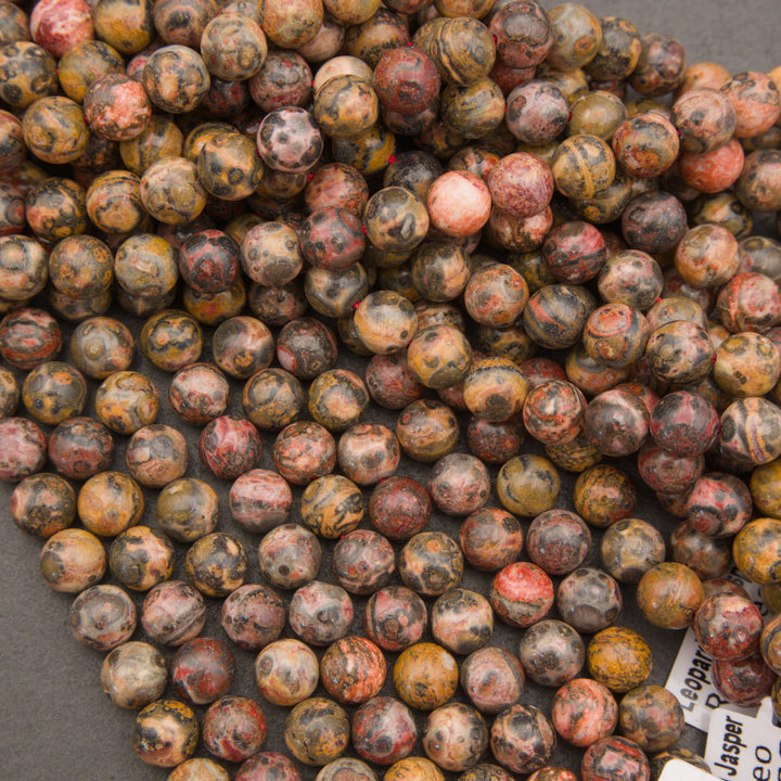 A pile of polished round gemstone beads in earthy marbled colors, including brown, gray, black, orange, and muted red, with a small white label visible in the lower right corner.