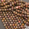 A pile of polished round gemstone beads in earthy marbled colors, including brown, gray, black, orange, and muted red, with a small white label visible in the lower right corner.