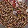 A pile of small round gemstone beads in earthy marbled colors, many tied with red string and attached to white barcode price tags, photographed on a gray surface.