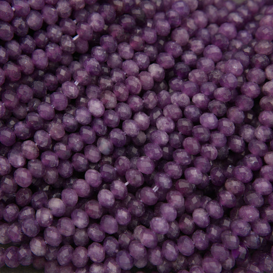A close-up pile of faceted purple beads, likely amethyst, showing a consistent round shape and polished crystal facets.