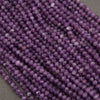 Multiple strands of faceted purple beads, likely amethyst, arranged diagonally on a neutral gray surface.