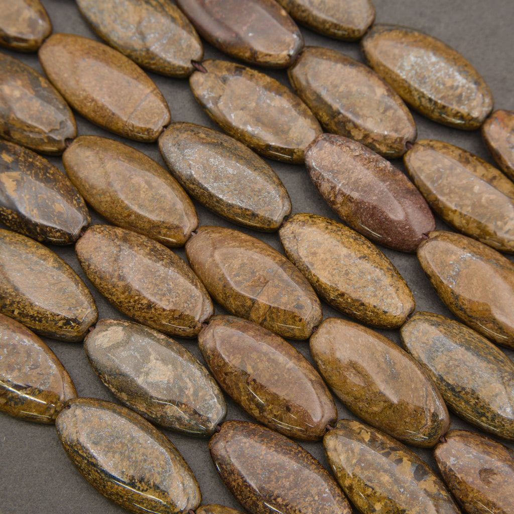 A close-up of polished oval beads in earthy brown, tan, and gold tones with natural marbled patterns, arranged diagonally on a dark gray background.