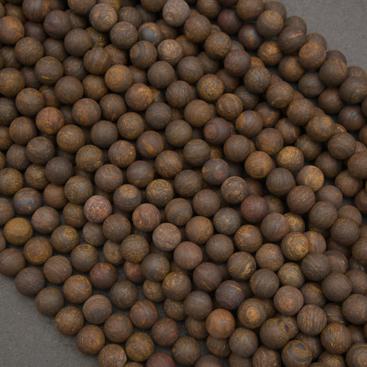 A close-up pile of small round brown beads with natural wood-like texture and subtle tan variation, photographed on a dark gray surface.