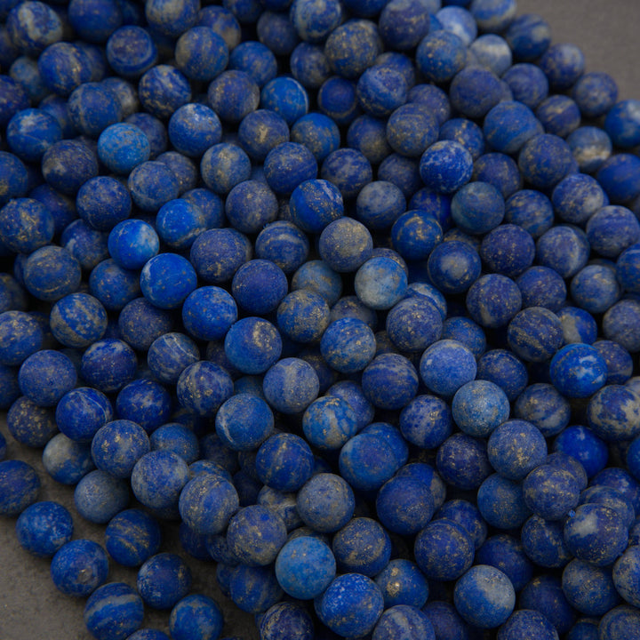 A pile of small round blue lapis lazuli stone beads with natural white and yellow veining, photographed close-up on a neutral gray surface.