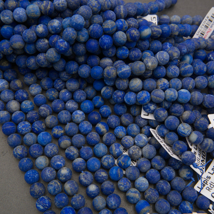 A pile of round blue lapis lazuli stone beads with natural white and light blue veining, shown on a neutral gray background with some white inventory tags visible.